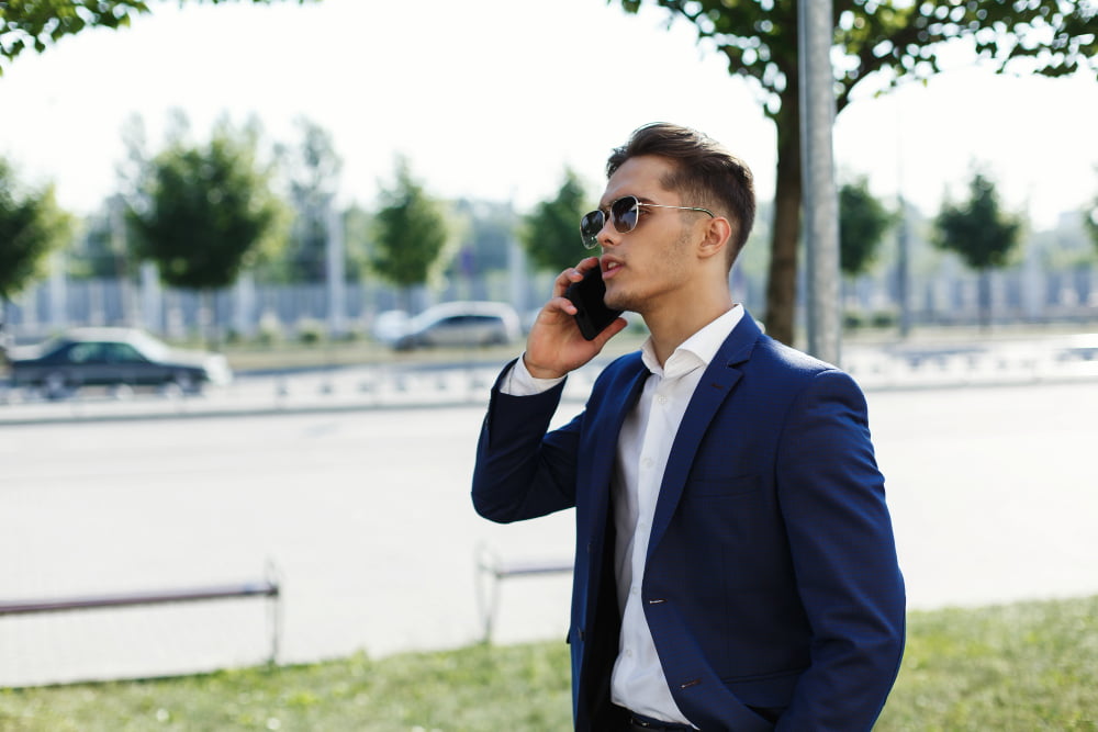 A man in a blue suit and sunglasses talks on his phone while standing outdoors near trees and a street in California.