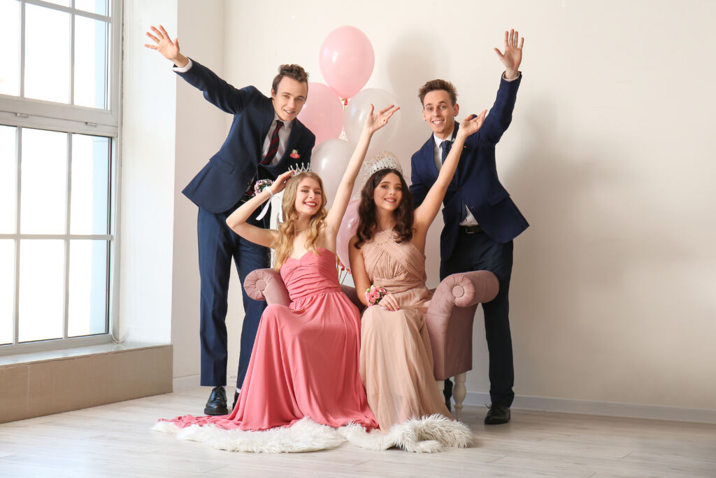 Two women in dresses and two men in suits from a local California suit outlet celebrate indoors with balloons, with the women wearing tiaras and smiling.