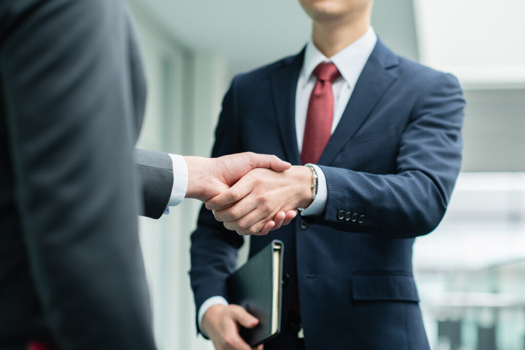 Two businessmen in suits shaking hands, one holding a folder, in a bright office setting.