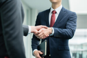 Two businessmen in suits shaking hands, one holding a folder, in a bright office setting.